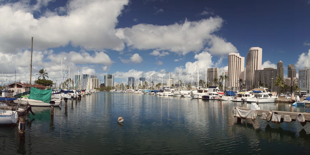 Sailboats and powerboats in their slips at the Ala Wai Small Boat Harbor on a clear day, Honolulu towers behind.